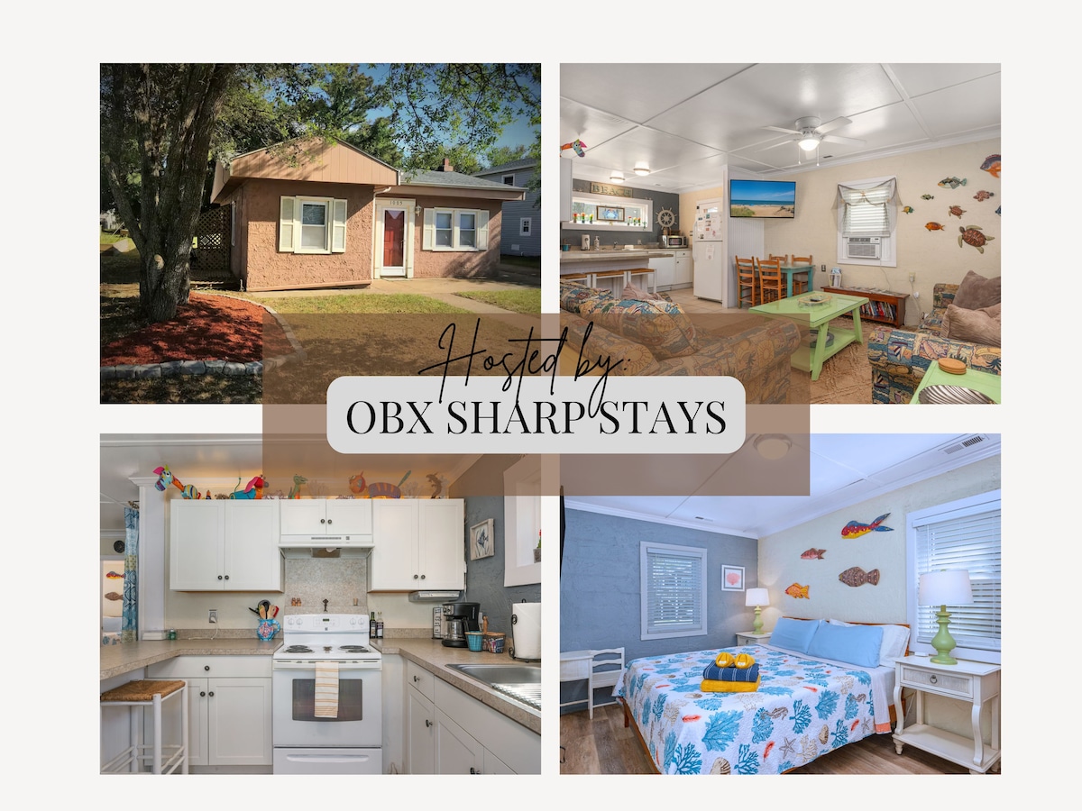 A charming cottage exterior is framed by lush greenery, featuring a welcoming front door and well-maintained landscaping. The interior showcases a cozy gathering space with light-colored furnishings, a dining area, and playful fish decor, creating a relaxed atmosphere for guests.