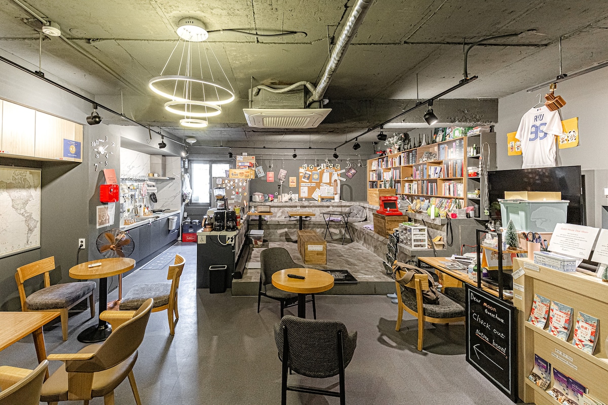 The communal lounge area features several wooden tables and chairs arranged for guest use. Shelving units display various books, while a relaxed seating area is centered on a soft rug. The space is illuminated by modern light fixtures and has a welcoming, open atmosphere.