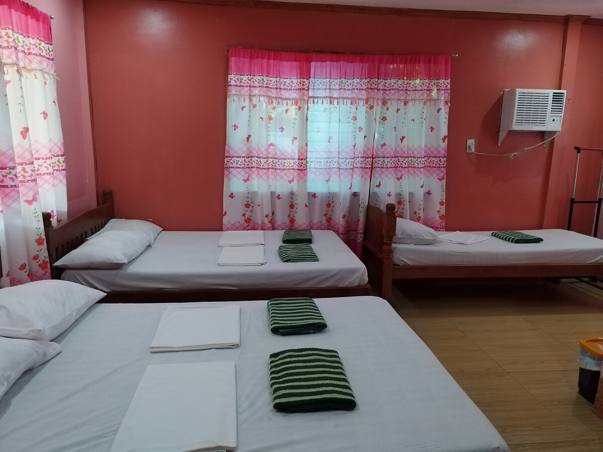 A bright room features three beds, each dressed with white linens and neatly arranged towels. Decorative curtains with a floral design frame a window, while an air conditioning unit is mounted on the wall. The wooden accents contribute to a cozy and welcoming environment.
