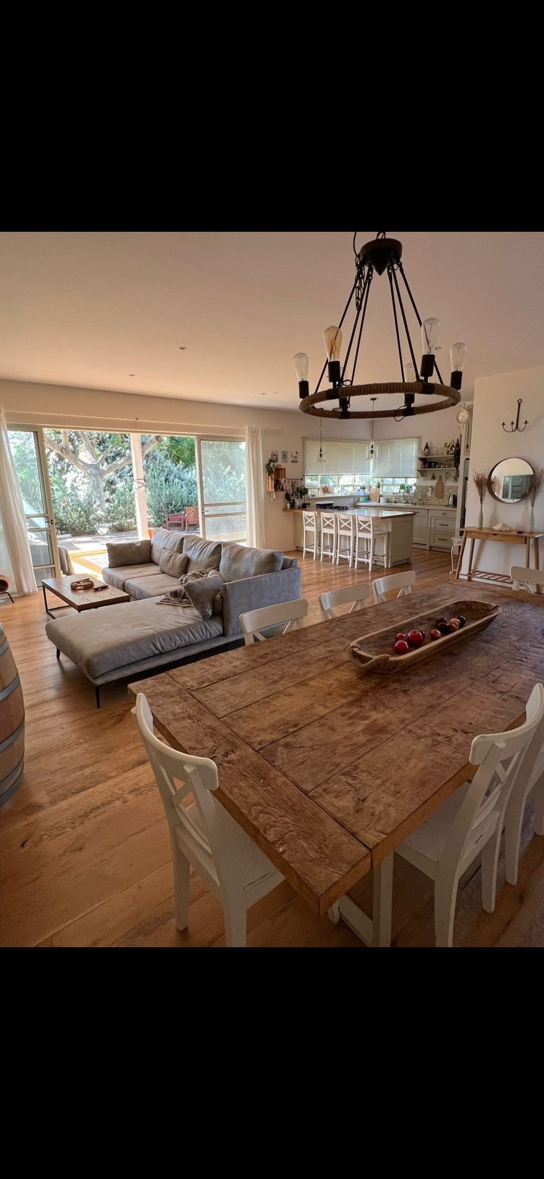 An inviting dining area features a rustic wooden table accompanied by white chairs. A cozy seating arrangement is positioned nearby, complemented by a large window that offers natural light and views of the outdoor greenery.