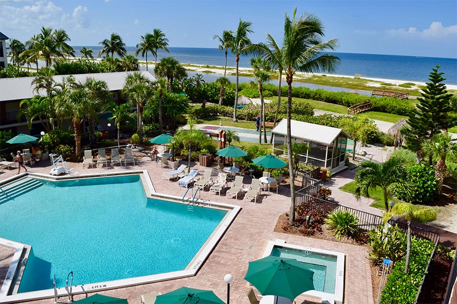 An aerial view captures the large pool and hot tub surrounded by lush greenery and palm trees. Sun loungers with green umbrellas are arranged around the pool area, while the beach and ocean are visible in the background under a clear blue sky.