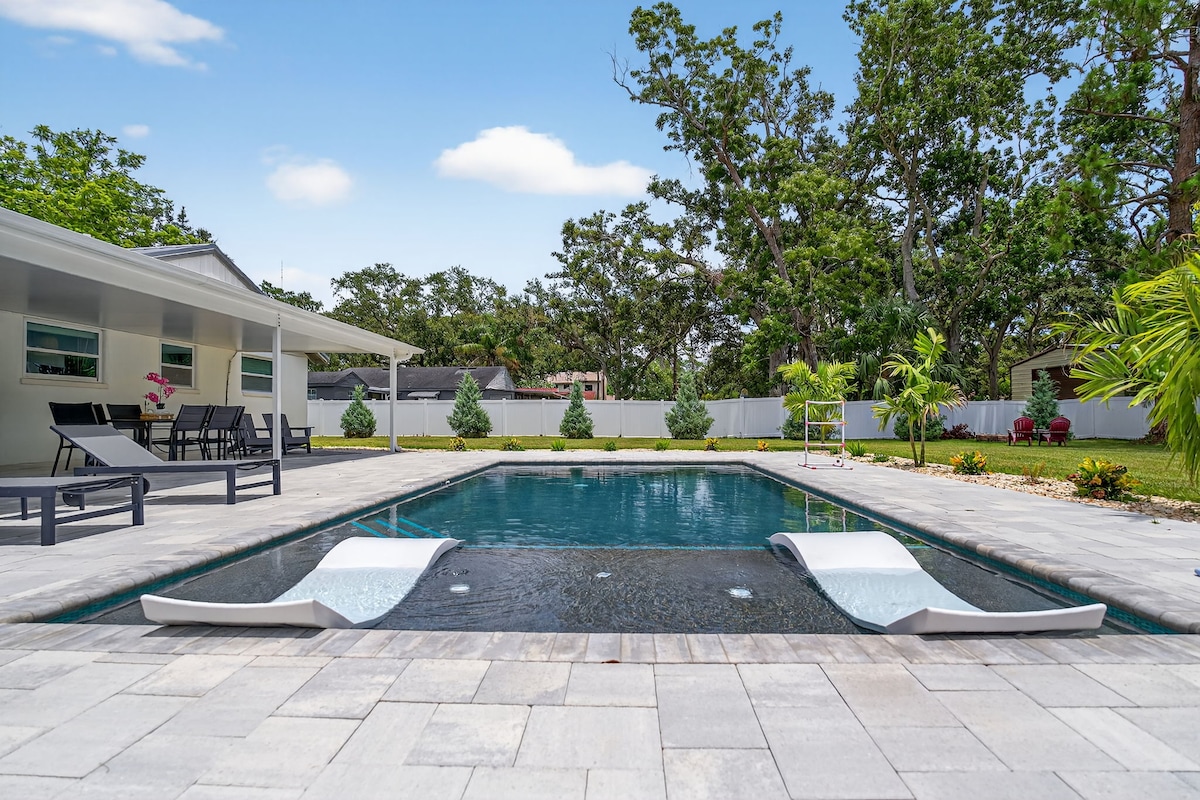 A newly designed backyard features a saltwater heated pool surrounded by spacious stone decking. Two loungers are positioned on the shallow Baja shelf, while greenery and landscaped bushes enhance the inviting space. A covered patio with seating is visible in the background.
