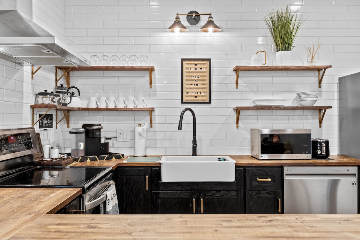 A modern kitchen is displayed with sleek black cabinetry and hardwood countertops. Open shelving holds dinnerware and glassware, while stainless steel appliances, including a dishwasher and microwave, are visible. A farmhouse-style sink is centrally located, with a stylish faucet above it.