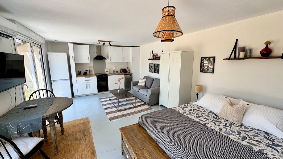 A spacious studio is presented with an open layout, featuring a cozy bed adorned with neutral bedding. A comfortable sofa sits nearby, accompanied by a dining table with two chairs. The kitchen is equipped with modern appliances, while ample natural light floods the room through large windows.
