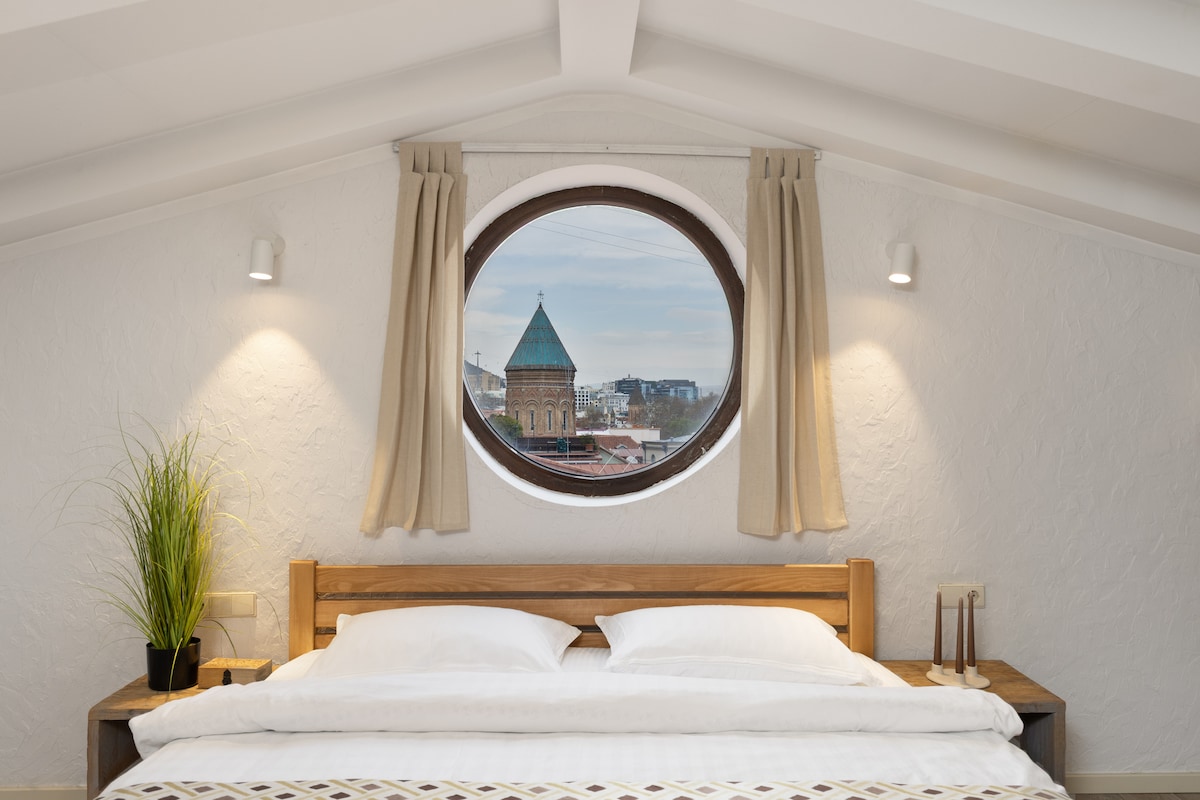 A cozy sleeping area features a spacious bed with white linens, complemented by a wooden headboard. A round window above frames a view of the city, with soft curtains on either side. Subtle wall lights enhance the warm ambiance of the room.