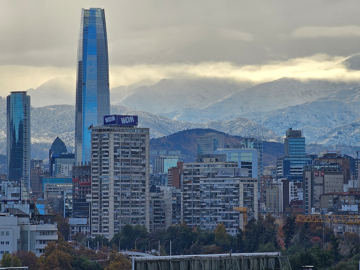 The image captures a panoramic view of an urban skyline, featuring a prominent skyscraper with a reflective glass facade. Snow-capped mountains are visible in the background, contrasting with the cityscape of modern buildings and greenery in the foreground.