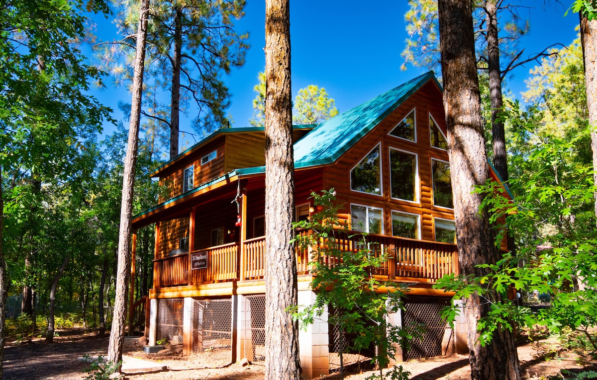 A log-style cabin is framed by tall trees, featuring a vibrant green roof and expansive windows. A spacious wrap-around porch is visible, inviting relaxation and outdoor enjoyment. Sunlight filters through the surrounding foliage, highlighting the natural beauty of the wooded lot.
