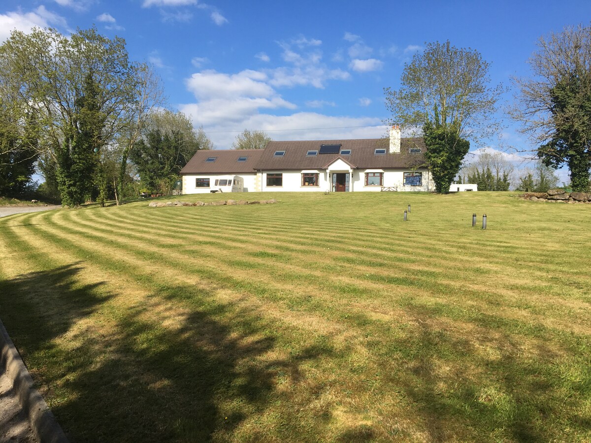 A single-story home is set in a spacious green lawn, featuring neatly arranged stripes of grass. The house is adorned with multiple windows, allowing ample natural light. Trees line the property, enhancing the serene countryside setting.