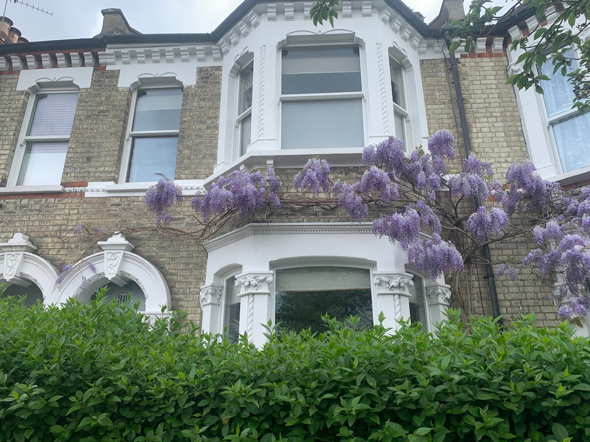 The exterior of a Victorian family home features a brick façade and tall windows. Delicate purple flowers cascade from a vine over the bay window, while a lush green hedge frames the front, enhancing the property’s charming character.