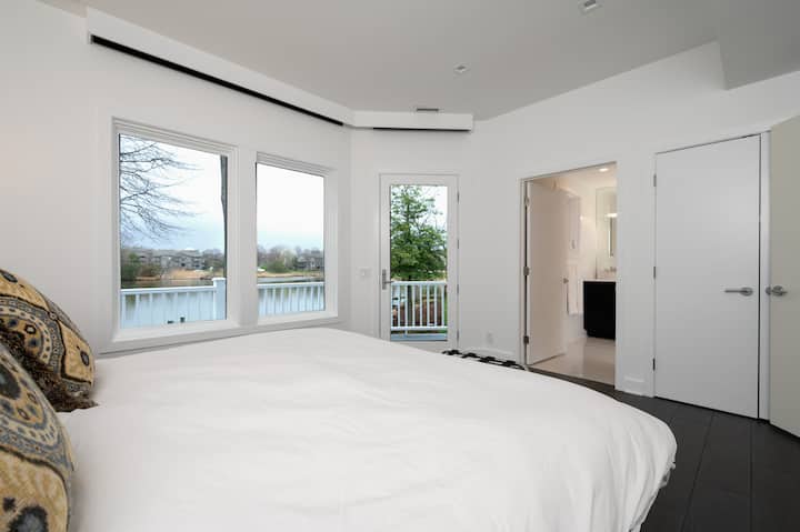 Dhoma e gjumit "king" për vizitorët në katin e poshtëm.  Krevati ka çarçafë dhe mbulesë me jorganë.  Poshtë jorganit. Hije elektronike të errëta për mbrëmjen.  Derë xhami në verandën e pasme.