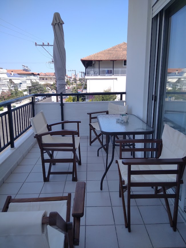 A balcony area features a glass-top table surrounded by four light-colored folding chairs. A sunshade is positioned for potential coverage, and the view includes nearby rooftops and clear blue skies. The tiled floor provides a clean, spacious area for relaxation.