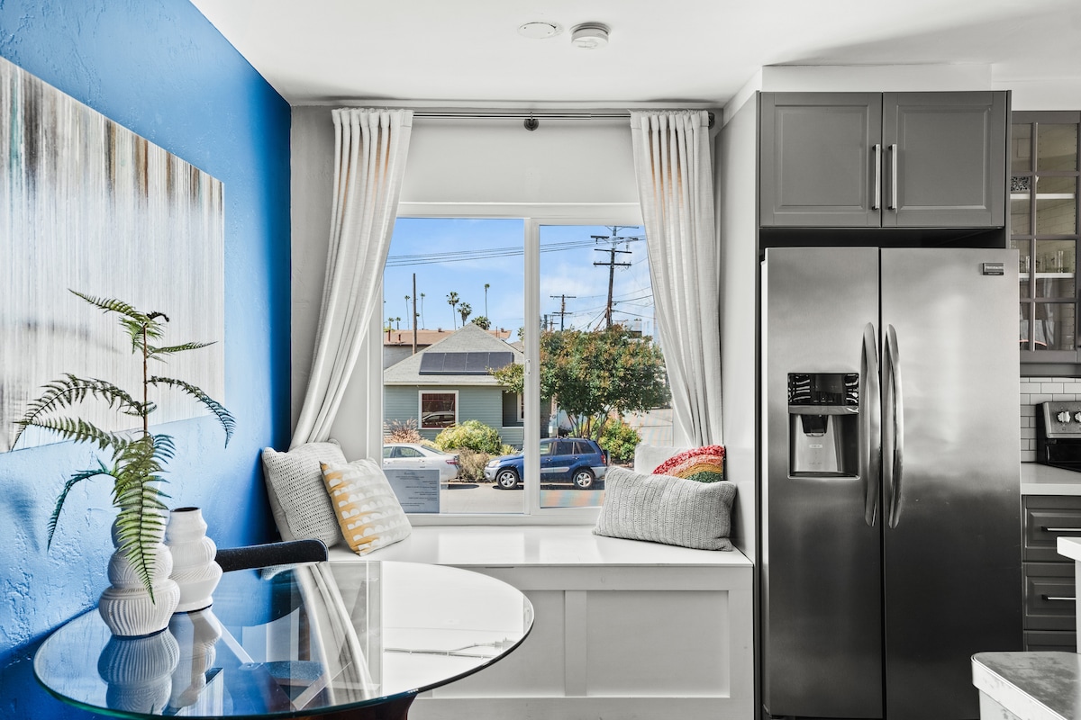 A bright kitchen area is highlighted by a blue accent wall and a large window overlooking the neighborhood. Cushions are placed on the window seat, inviting relaxation. A round glass table sits nearby, paired with a sleek stainless steel refrigerator.