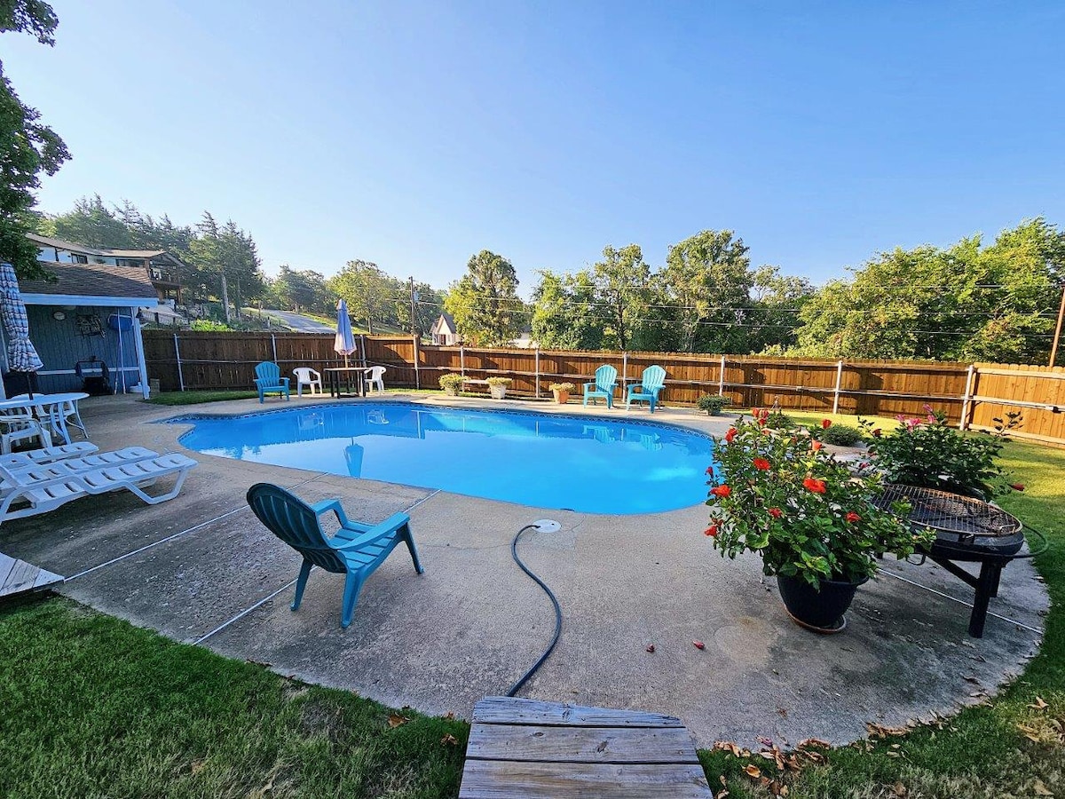 A spacious outdoor area features a swimming pool surrounded by a concrete deck. Colorful lounge chairs are positioned for relaxation, and vibrant flower arrangements add a touch of nature. A wooden fence encloses the space, ensuring privacy and comfort.