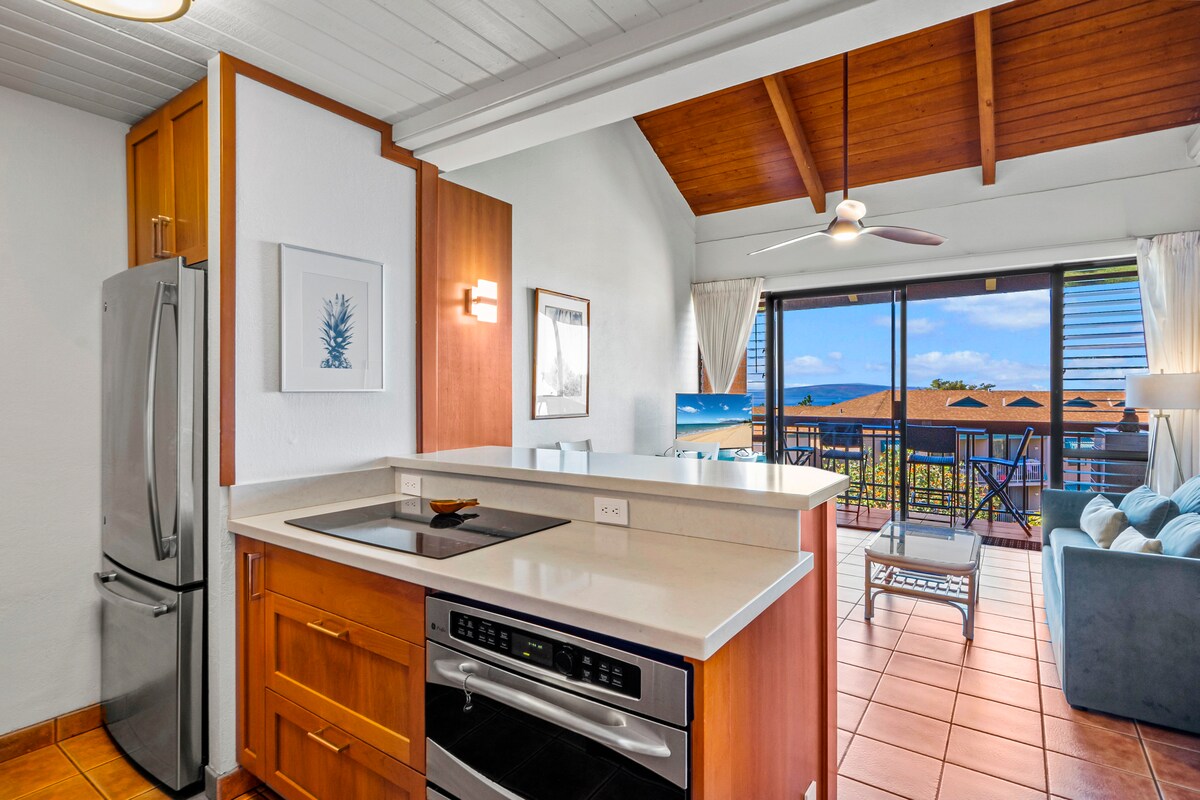 The kitchen area features modern appliances, including a stainless steel refrigerator and oven. A light-colored countertop offers ample workspace. High ceilings with wooden beams enhance the airy feel. Large windows provide views of the ocean and connect the indoor space to the outdoor balcony.