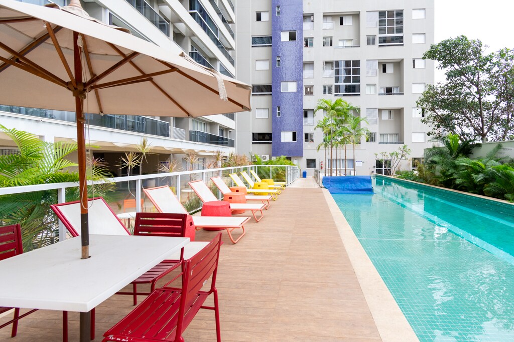 A spacious outdoor area is showcased with a long swimming pool reflecting the surrounding greenery. Colorful lounge chairs line the poolside, accompanied by a table shaded by a large umbrella. The sleek building stands in the background, enhancing the modern aesthetic.