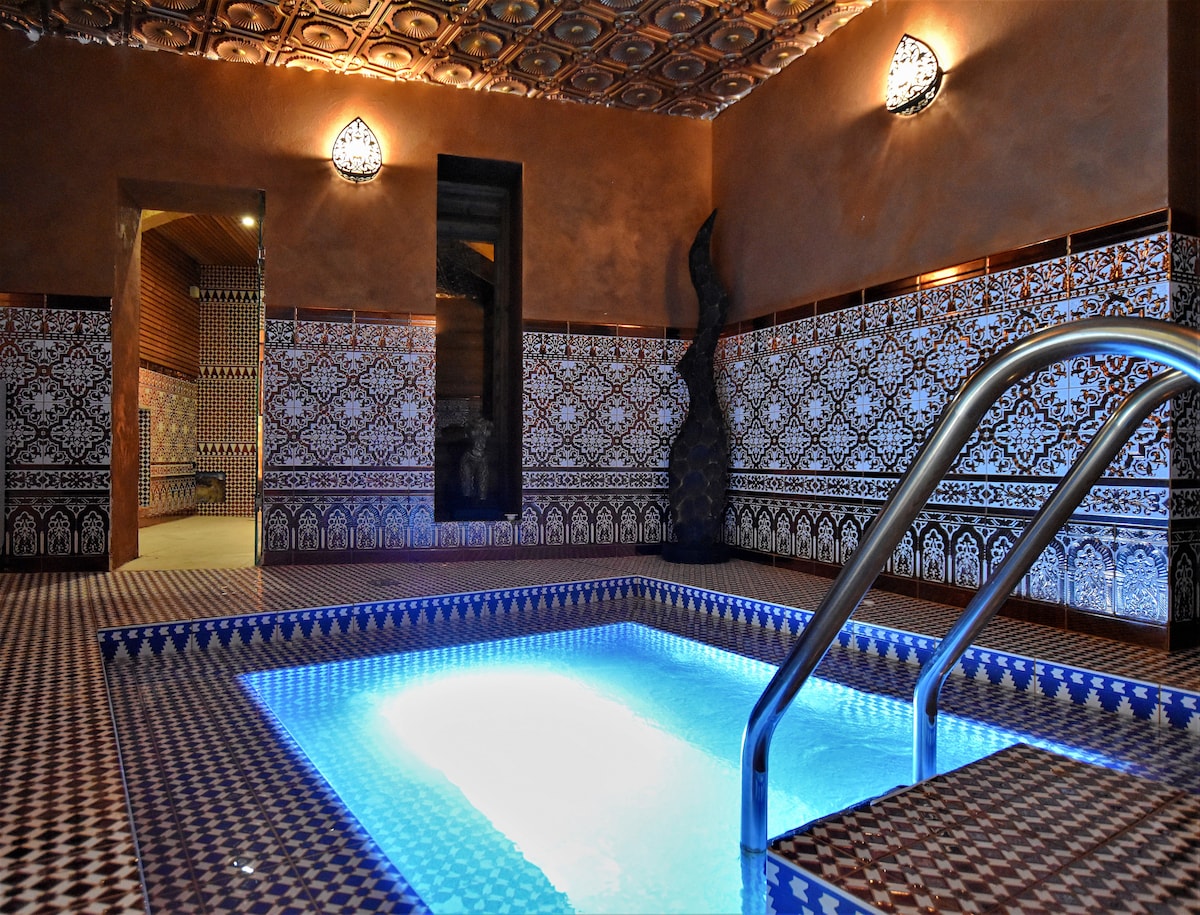 An indoor pool features intricate tile work in vibrant patterns along the walls and floor. The water is illuminated, creating a serene ambiance. Two ornamental lights are mounted on the walls, adding a subtle glow to the space.