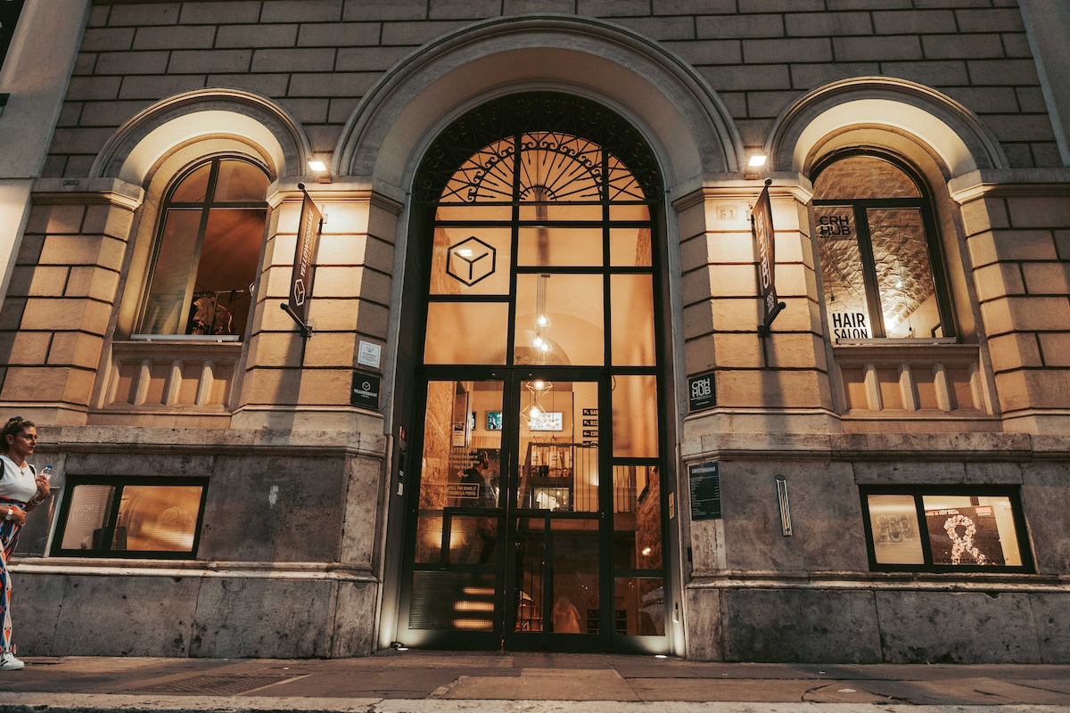 The entrance of the building is showcased, featuring large glass doors framed by elegantly arched windows. The facade displays intricate architectural details, and signage for the hair salon is visible. Soft lighting enhances the inviting atmosphere of the entrance.