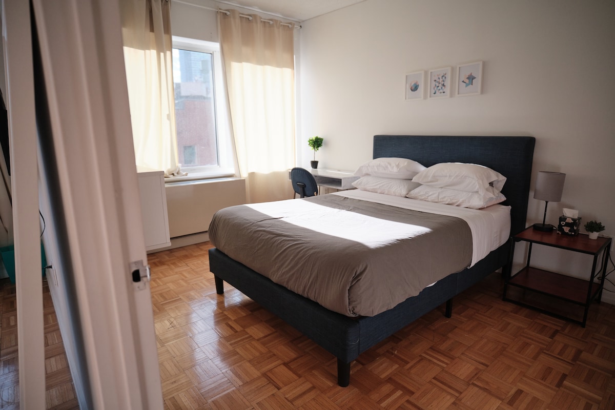 A well-lit bedroom features a spacious bed with layered bedding. Two windows are dressed with light curtains, allowing natural light to fill the room. A small desk is positioned beside the bed, complemented by a bedside table with a lamp.