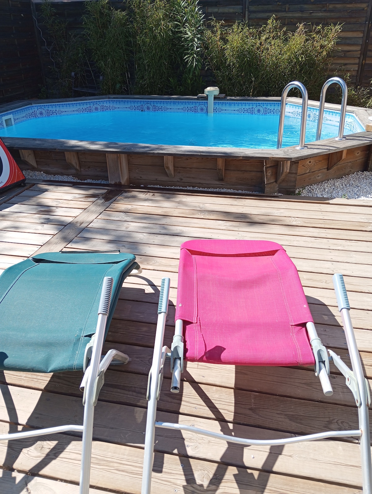 A small swimming pool is surrounded by wooden decking. Two lounge chairs, one green and one pink, are positioned elegantly near the water's edge. Lush bamboo plants provide a natural backdrop, enhancing the serene outdoor setting.