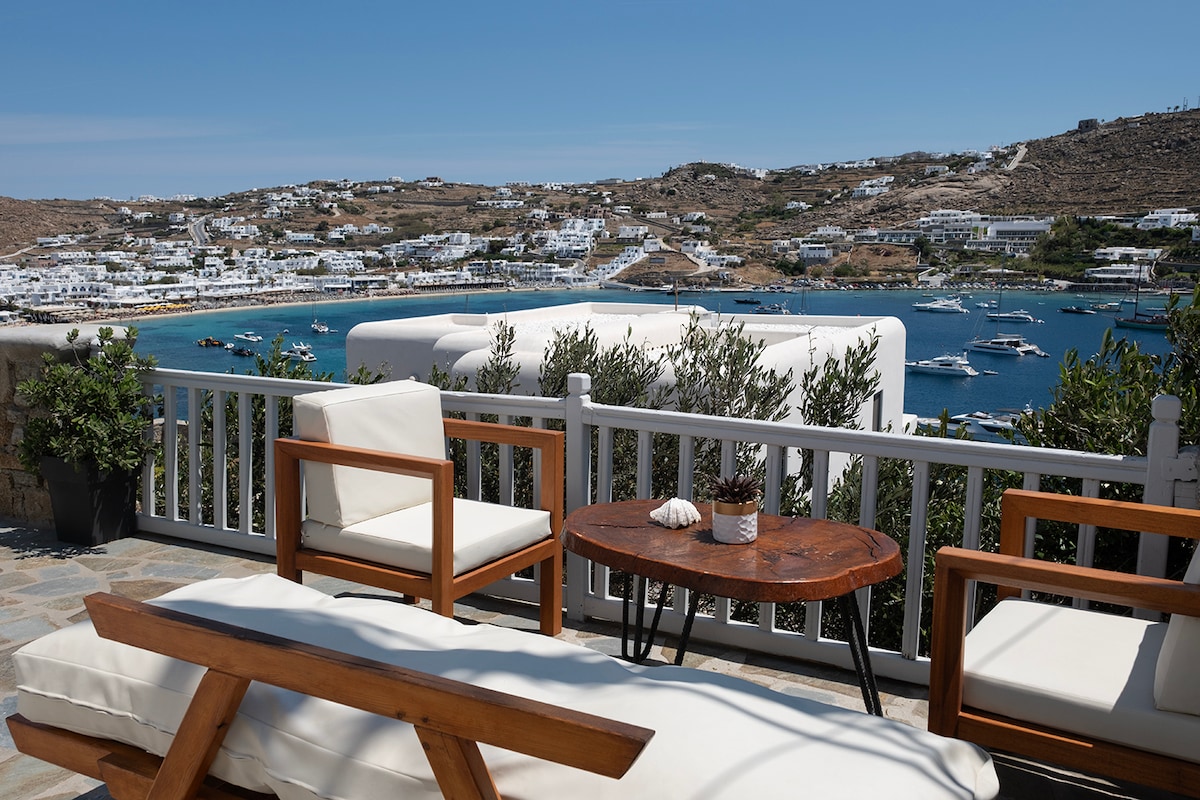 A serene outdoor seating area is showcased, featuring two wooden chairs and a small round table. A scenic view of the coastline, dotted with boats, is visible in the background, framed by lush greenery and clear blue skies.