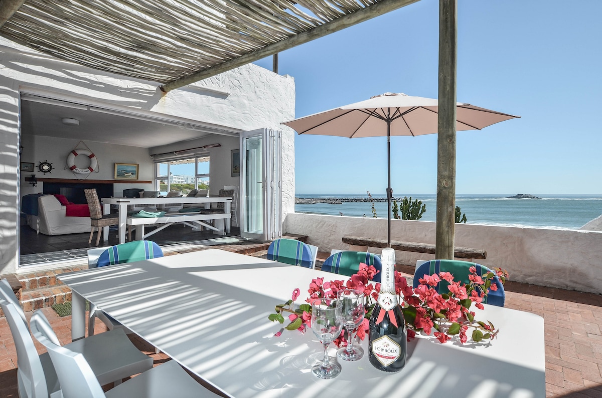 An outdoor dining area is furnished with a white table set for four, adorned with pink flowers and a sparkling wine bottle. A shade umbrella offers protection from the sun, and ocean views stretch in the background, providing a serene backdrop for meals.