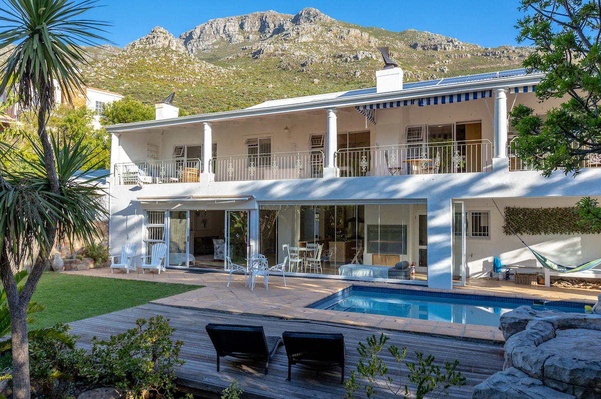 The exterior of the home showcases a spacious patio area adjacent to the swimming pool, flanked by manicured lawns. Sun loungers are positioned near the pool, with a mountain backdrop enhancing the tranquil setting. Large glass doors allow seamless access to indoor living spaces.