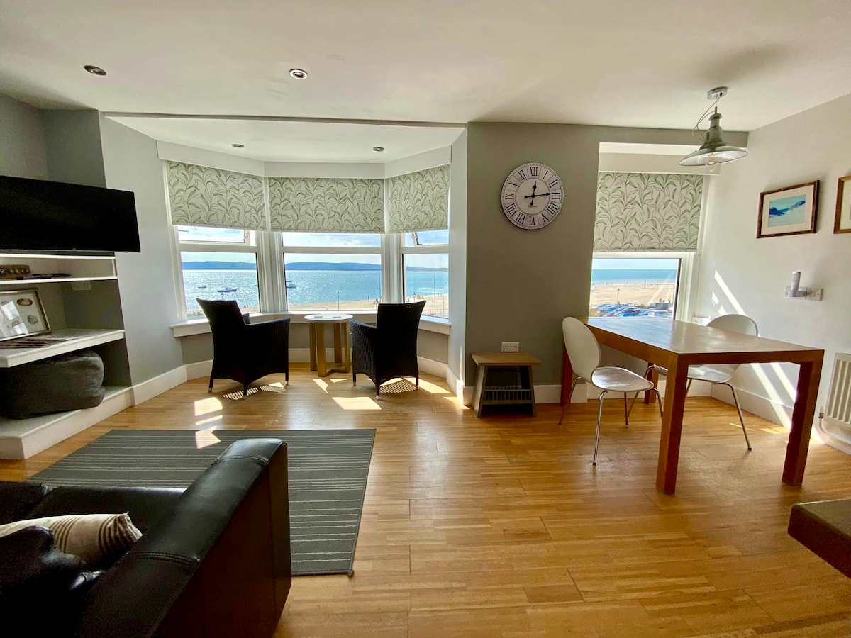The living area features large windows with sea views, allowing natural light to fill the space. A comfortable black sofa is positioned nearby, while a dining table and chairs are arranged for dining. Decorative elements include a wall clock and artwork, enhancing the atmosphere.