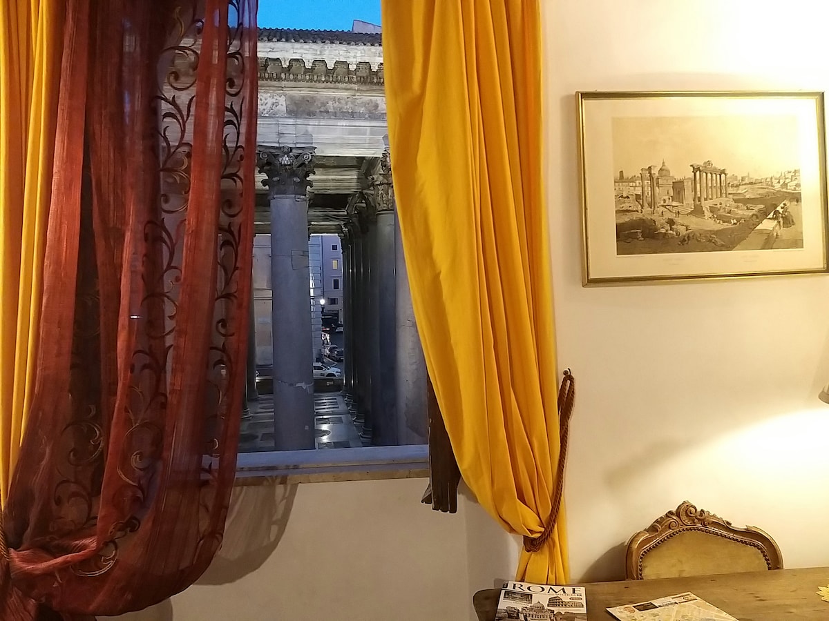 A window view reveals the Pantheon beyond flowing curtains in rich maroon and gold. An antique-style sitting area is positioned nearby, featuring a detailed chair and a small table with informational materials about Rome. Soft lighting from a lamp enhances the cozy ambiance.