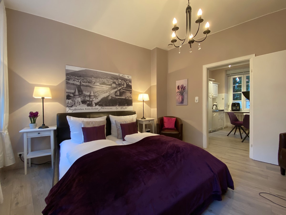 A spacious room features a large bed covered with a soft purple blanket. A stylish chandelier hangs above, and a bedside lamp provides warm light. A picturesque wall art depicting a landscape is displayed above the bed, while an open doorway leads to a kitchenette area.
