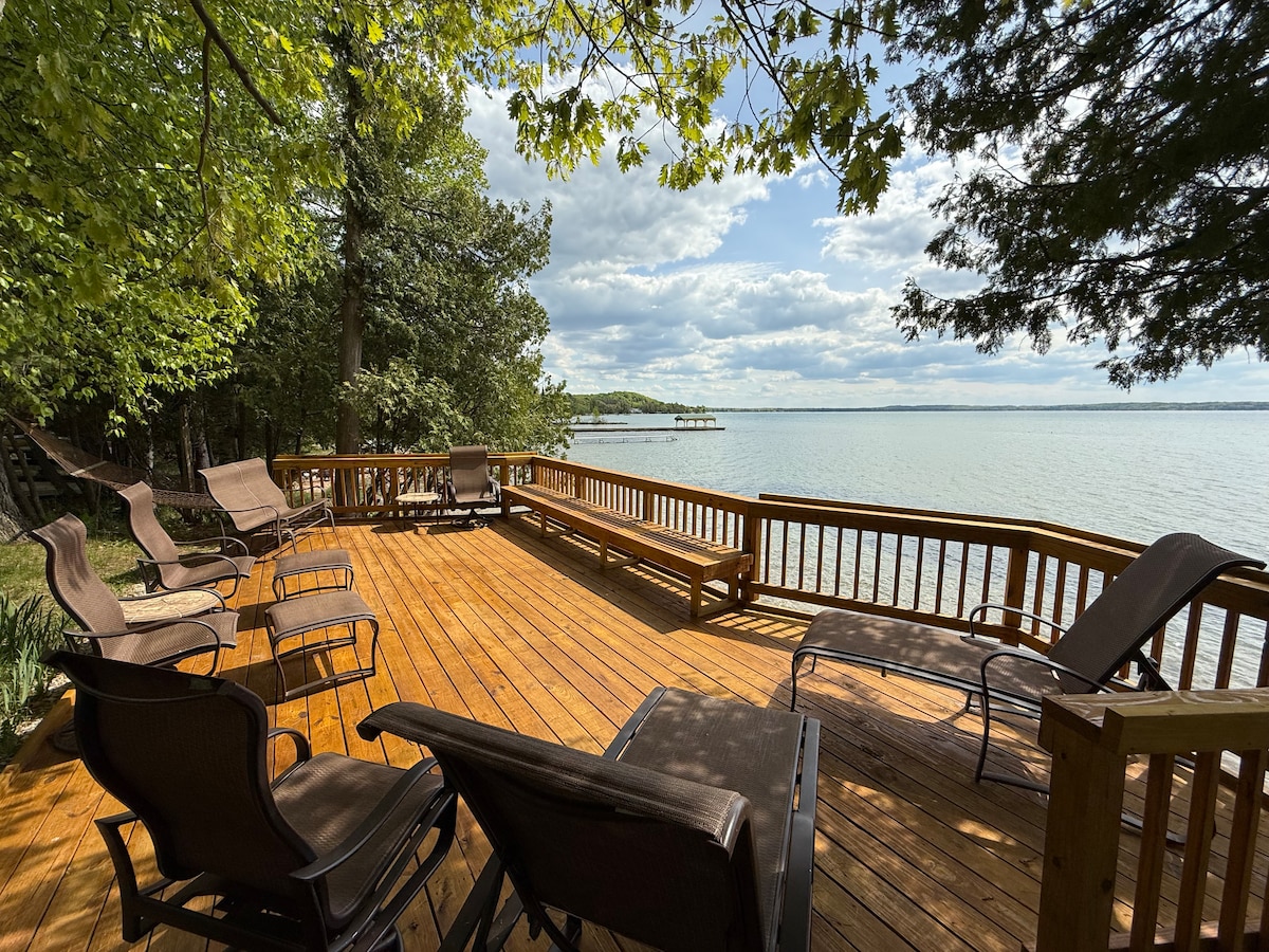 A spacious wooden deck overlooking Torch Lake is surrounded by trees. Sun loungers are arranged for relaxation, offering views of the calm water. The deck extends toward the horizon, providing an inviting area for outdoor activities and enjoying the natural surroundings.