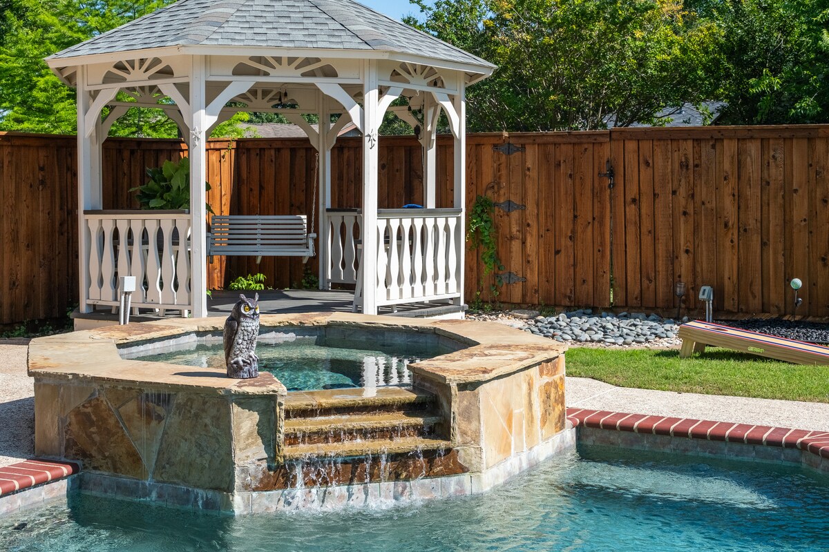 A charming gazebo is positioned within a landscaped backyard, featuring a hot tub and a small waterfall. A bench swing hangs in the gazebo, surrounded by green foliage, with a wooden fence providing privacy. A lounge chair is visible beside the swimming pool.