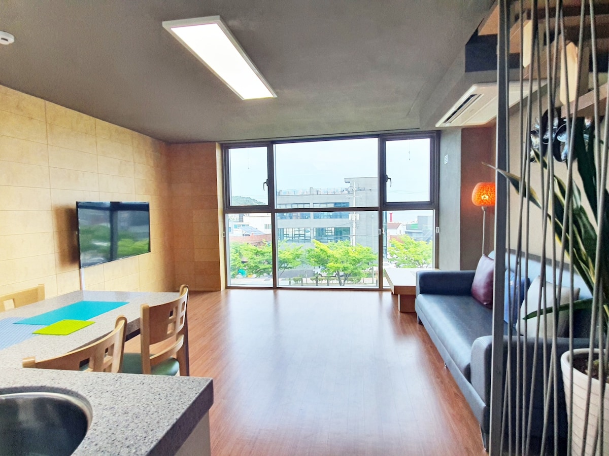 The spacious living area features large windows, providing ample natural light and a view of the city. A comfortable sofa with pillows is positioned beside a dining table, while a mounted television is visible on the wall, enhancing the modern design of the interior.