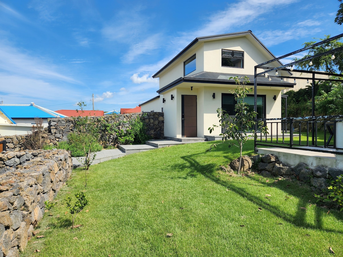 A two-story house is surrounded by a spacious green lawn. A stone wall encircles part of the property, and a pathway leads to the entrance. The building features large windows and a modern design, complemented by clear blue skies above.