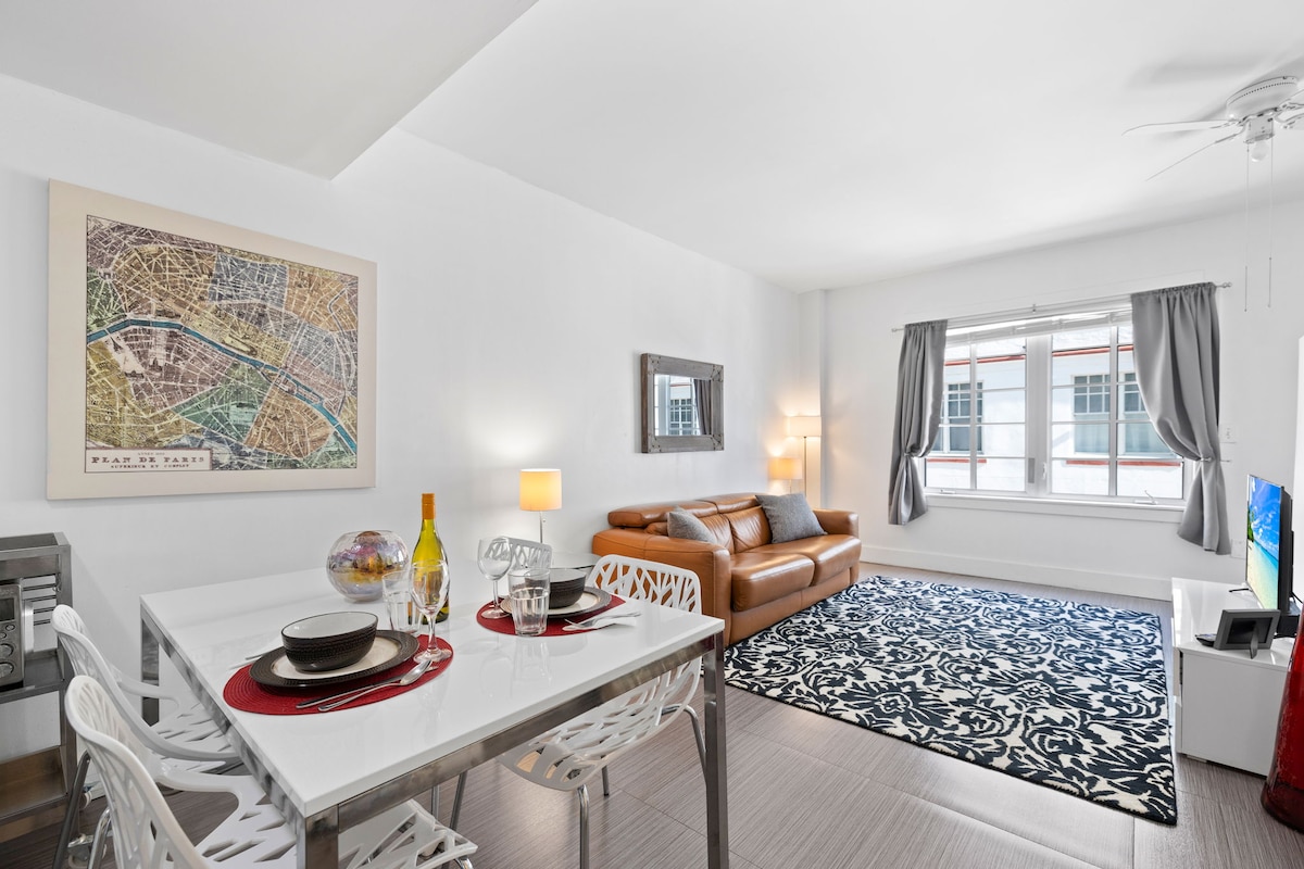 A bright living space features a modern dining table set for two, complemented by a colorful map artwork on the wall. A spacious leather sofa is positioned opposite a television, with light filtering through large windows adorned with sheer curtains.