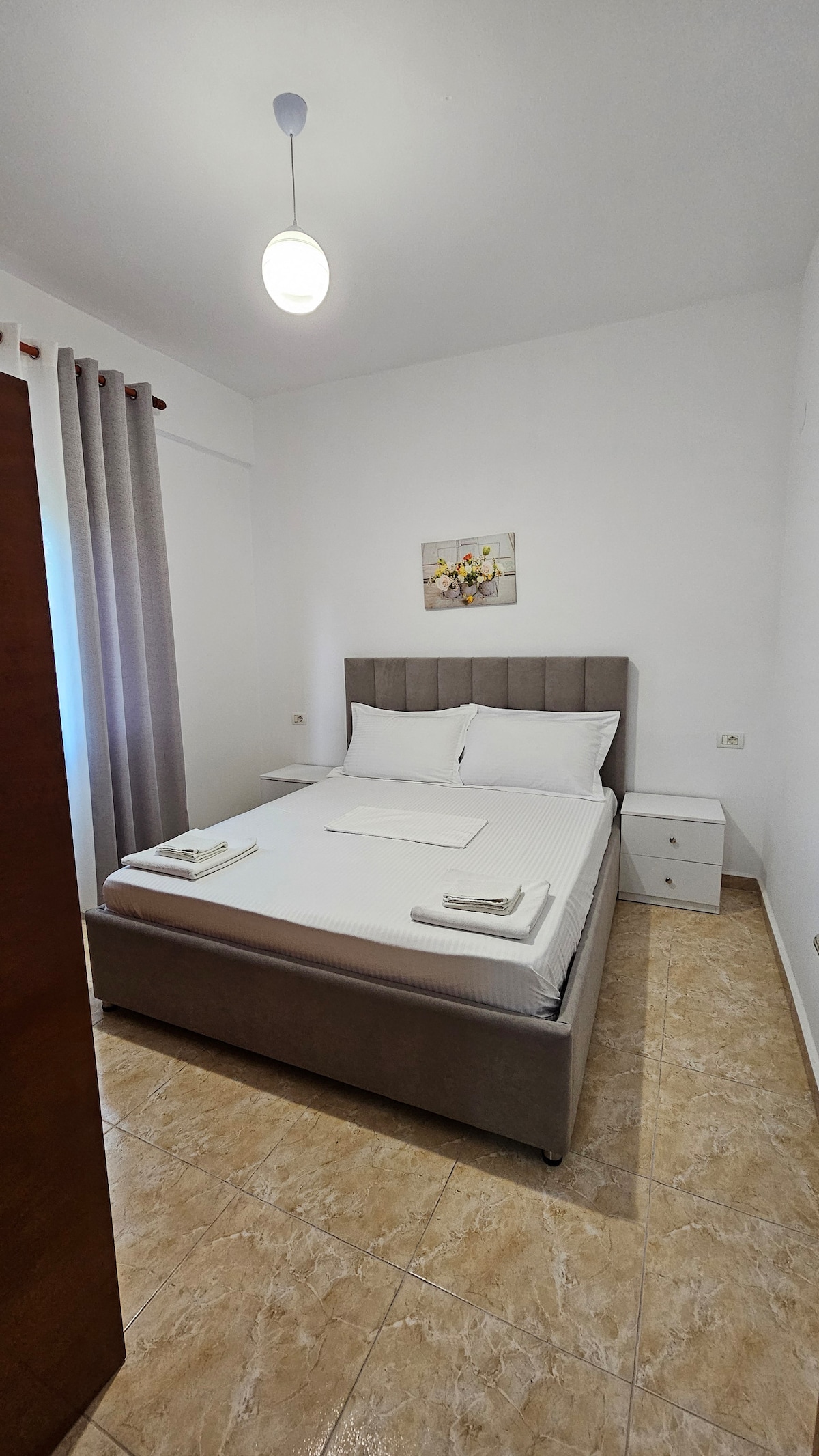 A neatly arranged bedroom features a cozy bed framed by soft, neutral-toned walls. A bedside table provides space for personal items, while a floral artwork adds a subtle touch of decoration. Light filters in through the window, complemented by pale curtains.