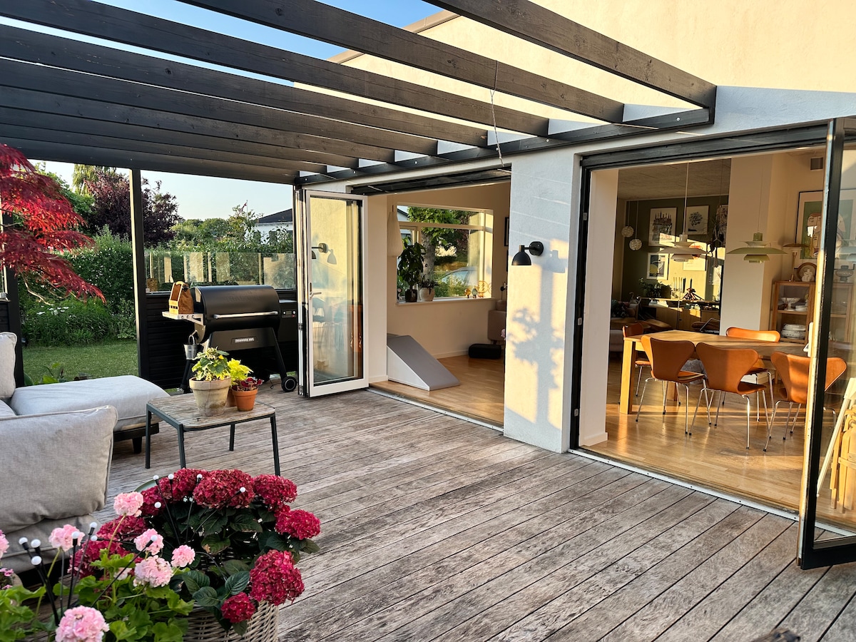A spacious terrace features a wooden deck, complemented by a seating area with outdoor sofas and potted flowers. Large glass doors open towards a well-lit indoor dining space with a table and chairs. A grill is positioned nearby, surrounded by greenery.