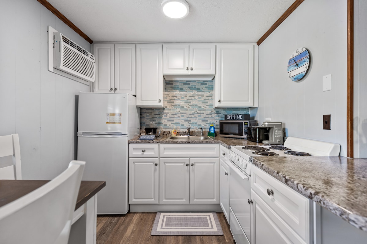 The kitchenette features bright white cabinetry and granite countertops, equipped with a refrigerator, microwave, oven, coffee maker, and toaster. A tiled backsplash adds a touch of style. A small dining table is visible, providing a cozy dining space.