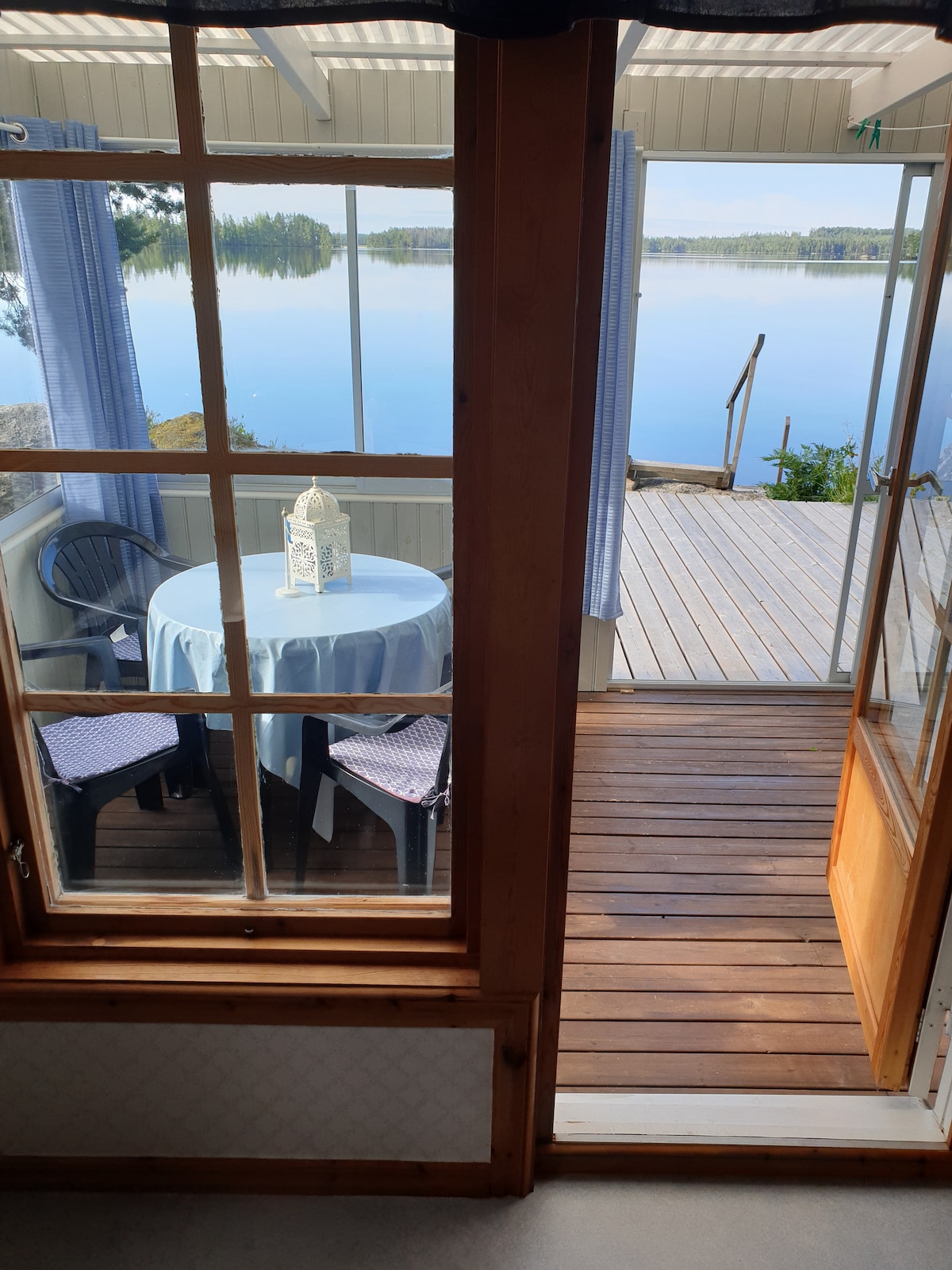 A view from inside the cabin showcases a glass door leading to an outdoor deck. A round table with a light blue tablecloth is set with matching chairs, while the calm lake is visible through large window panes, adding a serene backdrop.
