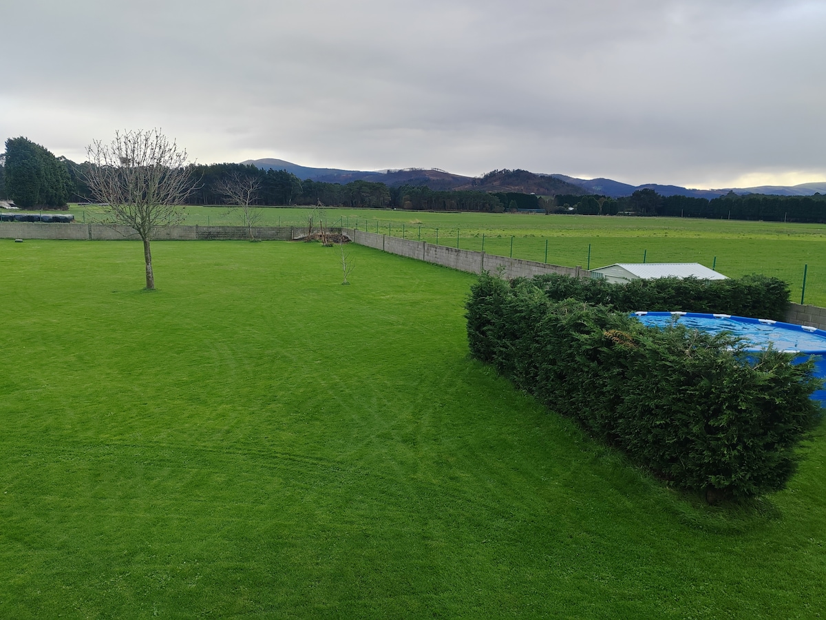A spacious green lawn stretches across the area, bordered by a low hedge. In the background, rolling hills are visible under a soft, cloudy sky. A pool is situated toward one side of the yard, surrounded by a protective fence.