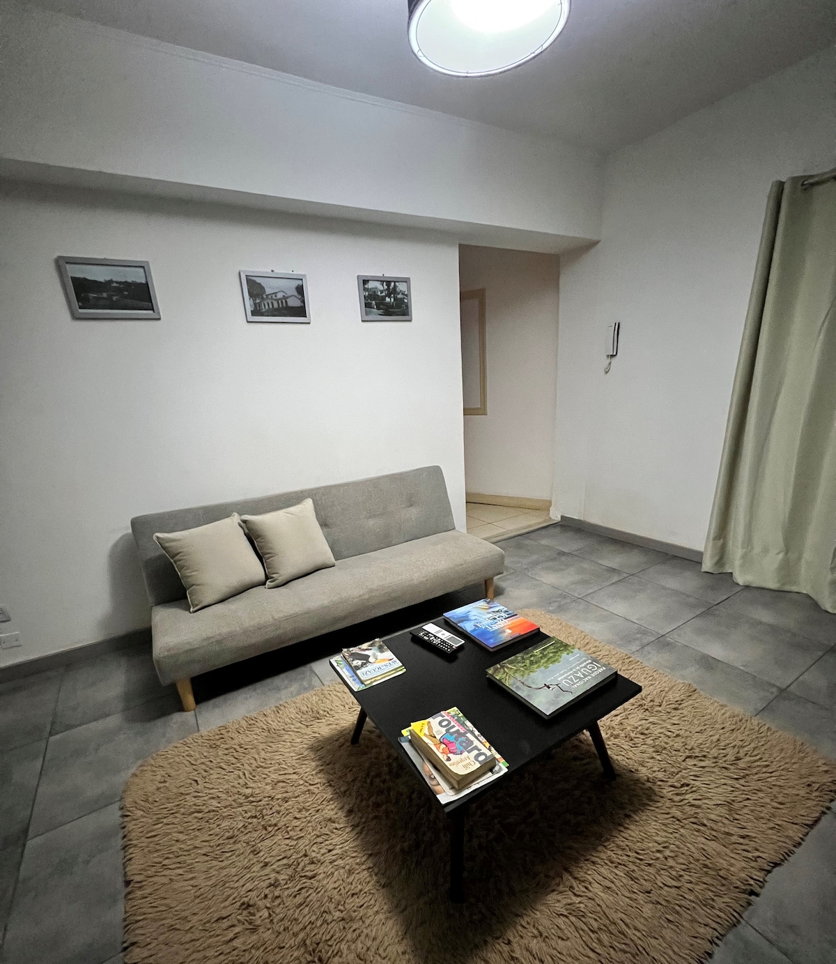 A cozy sitting area features a gray futon with two decorative pillows, paired with a low black table displaying various magazines and books. The floor is covered with a light-colored rug, and framed black-and-white photographs adorn the white walls.