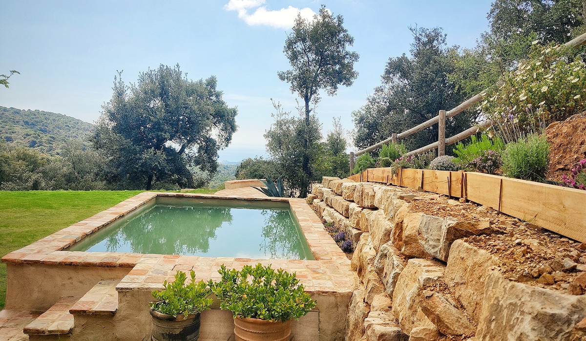 A private pool is set within a natural stone terrace, surrounded by greenery and flowering plants. A grassy area extends towards gentle hills in the background, providing a tranquil atmosphere. Wooden fencing complements the landscape, enhancing the connection with the serene outdoor environment.
