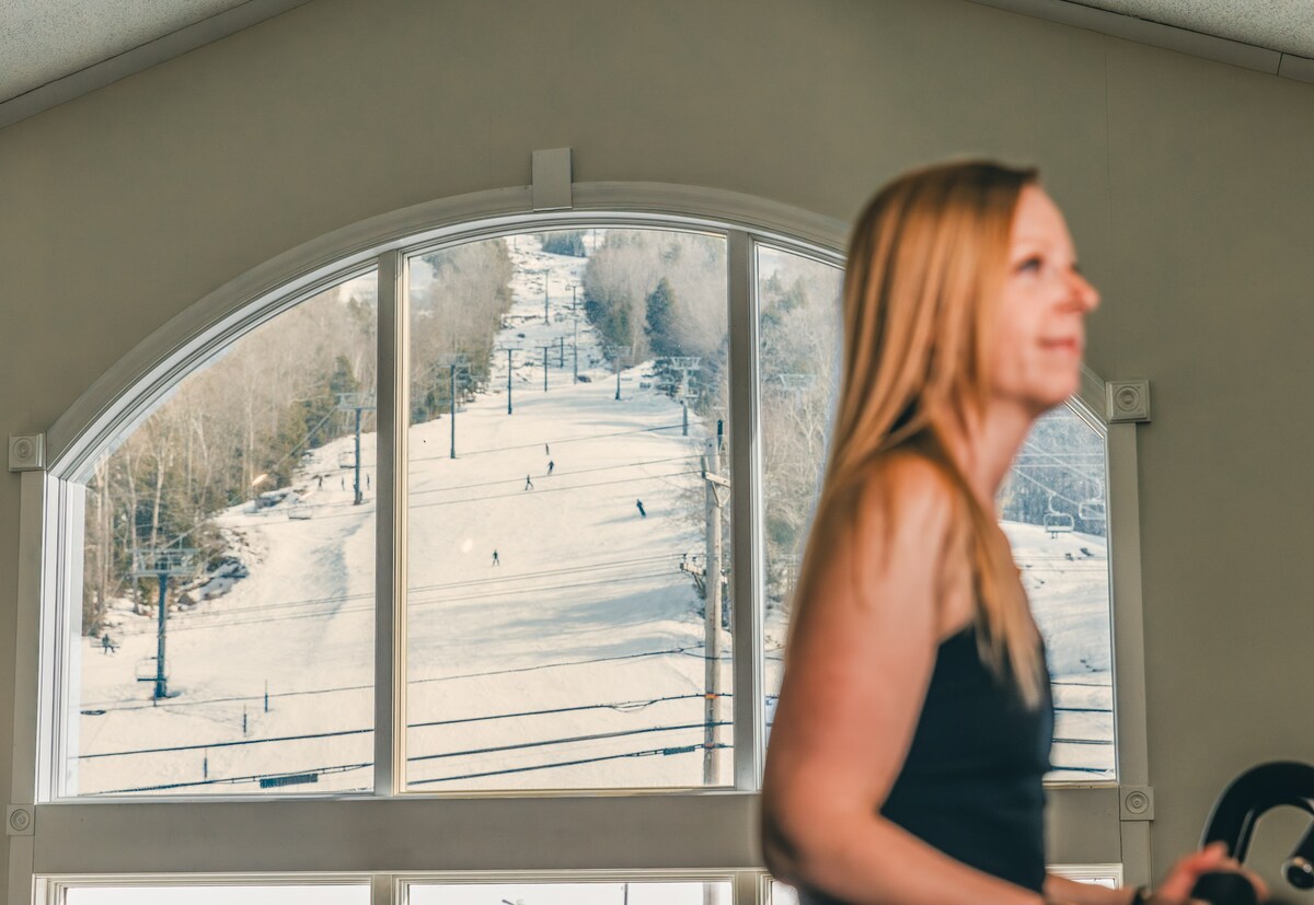 A large arched window frames a snowy landscape, revealing ski slopes dotted with skiers. The interior features a workout area, with a person engaged in exercise, suggesting an active lifestyle amidst the scenic winter backdrop.