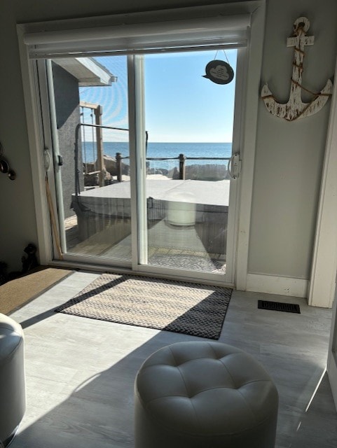 A view through glass sliding doors captures a serene lake scene. Sunlight filters in, illuminating a light-colored rug and a cozy round ottoman. Outside, a hot tub is positioned on the deck, framed by railings, offering a glimpse of the water beyond.
