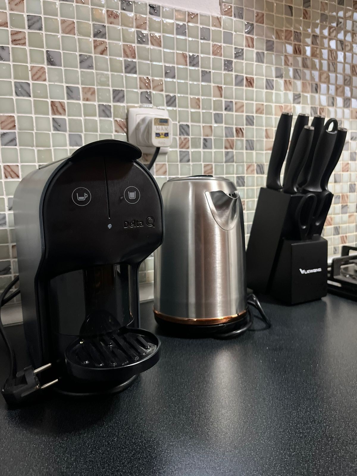 A modern kitchenette is illustrated, featuring a coffee maker and a stainless-steel kettle positioned beside a set of knives housed in a black block. The countertop has a smooth finish, and a mosaic tile backsplash adds texture to the setting.