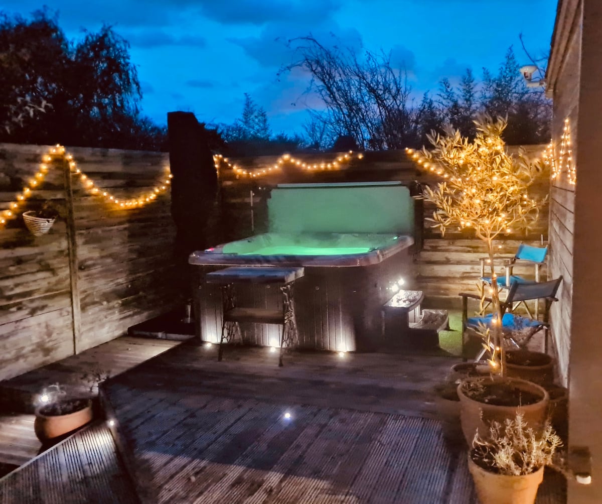 A private hot tub is surrounded by softly illuminated plants and string lights in the evening. Wooden decking offers a natural feel, while planters add greenery. The tranquil setting outside the lodge invites relaxation under a starlit sky.