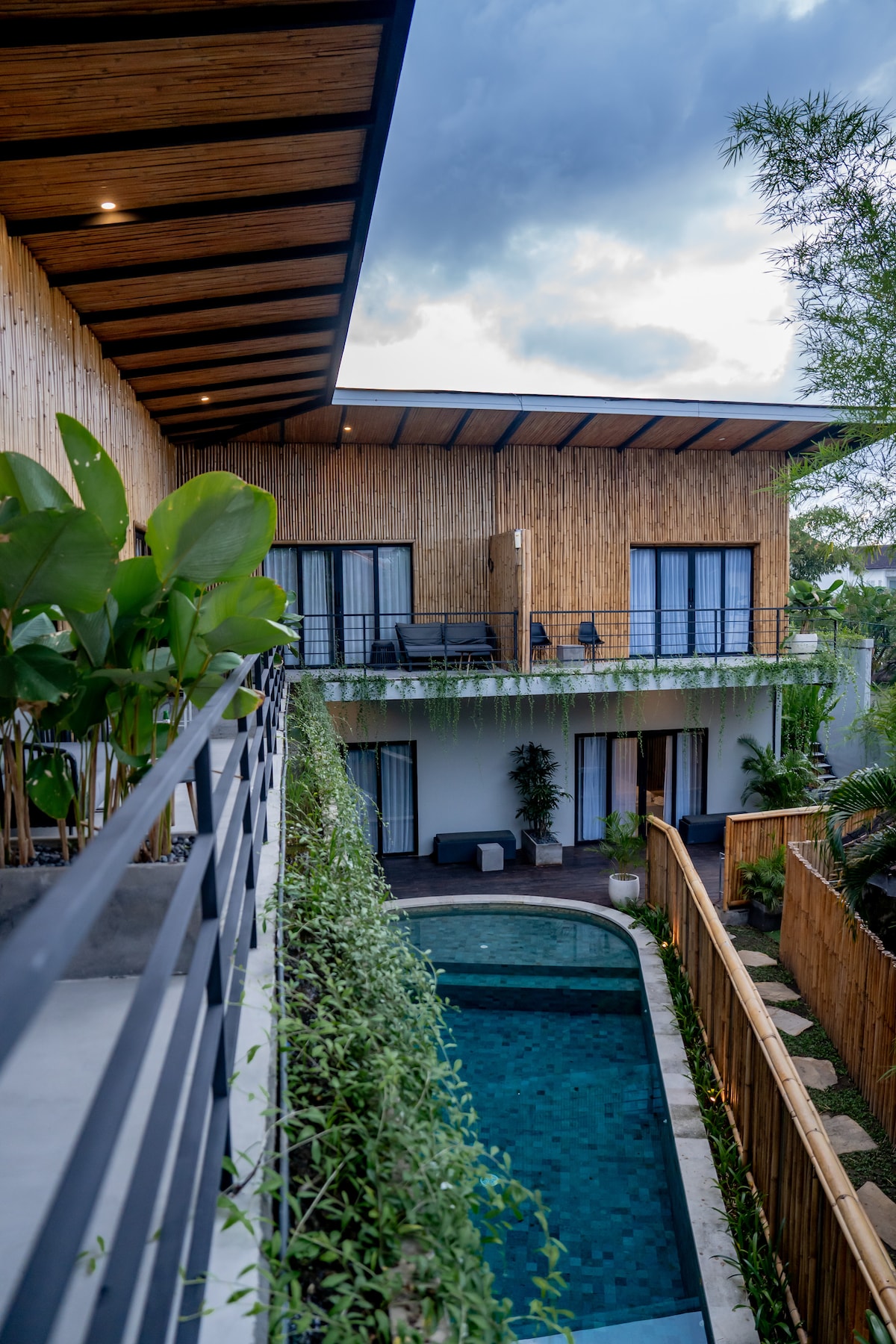 A modern building with natural wood accents is depicted, showcasing a linear design. A swimming pool is visible below, surrounded by lush greenery. The balconies above feature black railings and chairs, while large windows provide glimpses into the interior spaces.