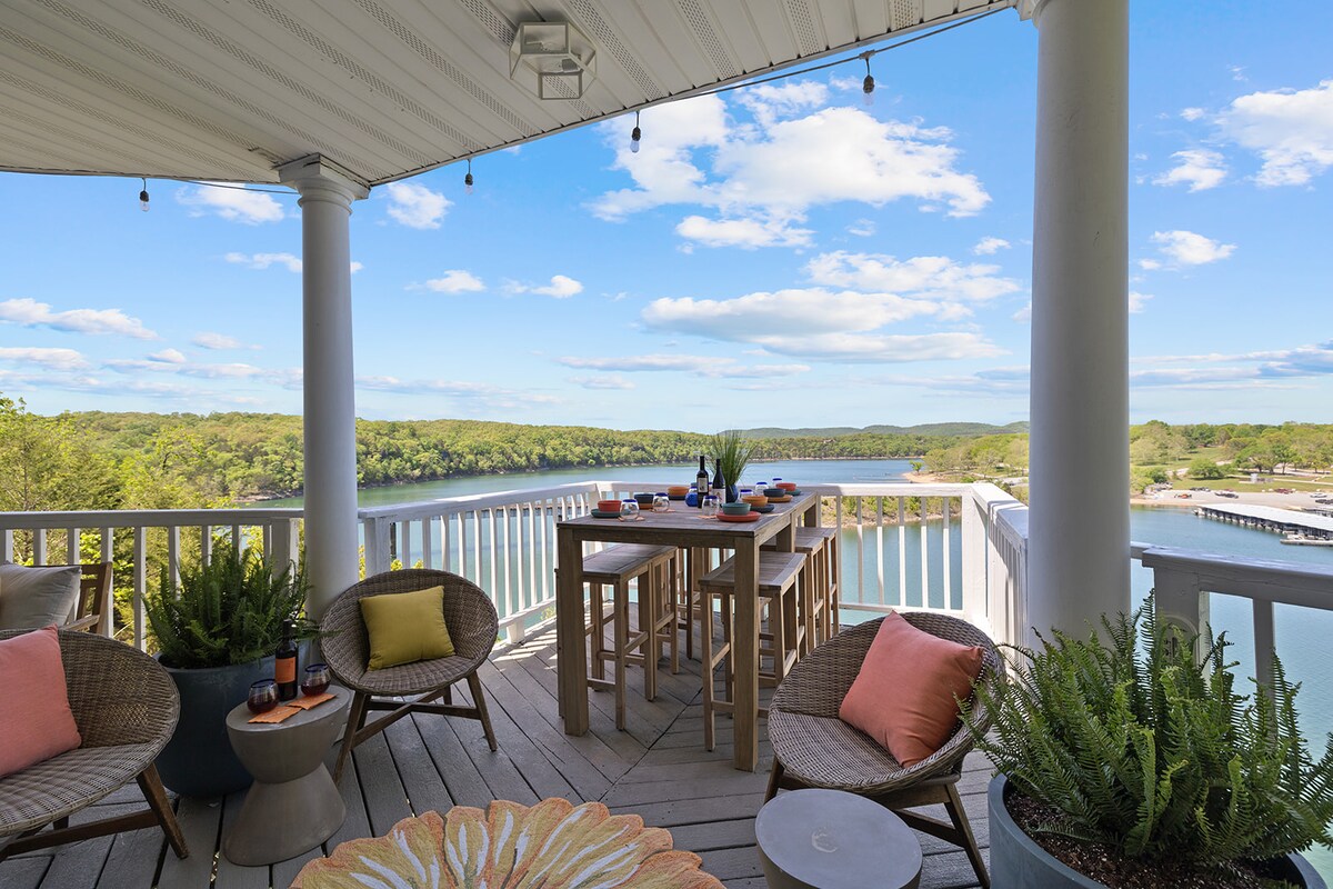 A spacious balcony features stylish outdoor seating and a large dining table. The area is surrounded by green trees and offers an unobstructed view of Table Rock Lake. Natural light brightens the space, creating an inviting atmosphere for outdoor gatherings.