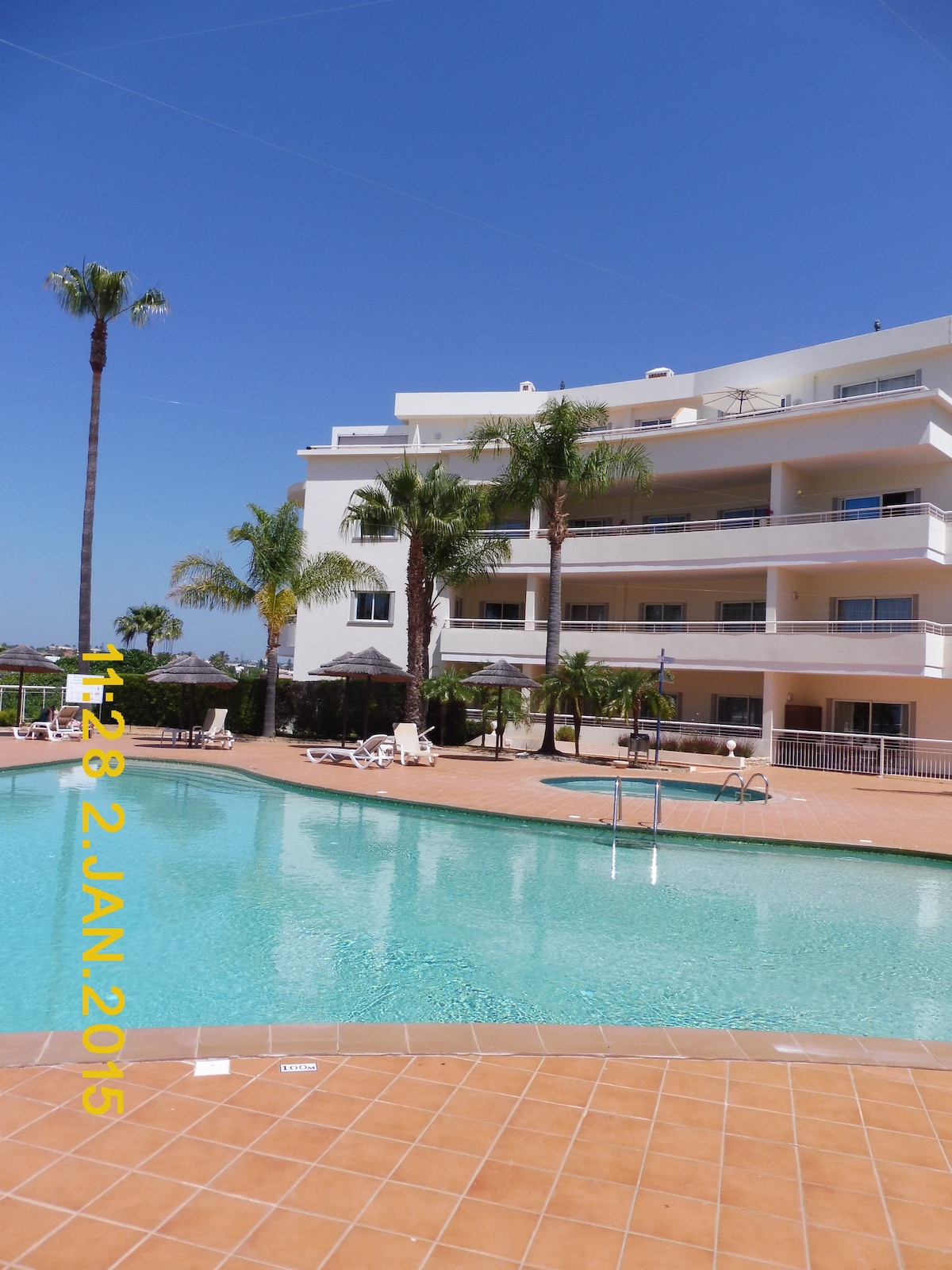 The image captures a modern multi-story building surrounded by palm trees, facing a clear blue sky. A spacious swimming pool is visible in the foreground, with lounge chairs positioned nearby. The pool area is complemented by tiled deck and green landscaping.