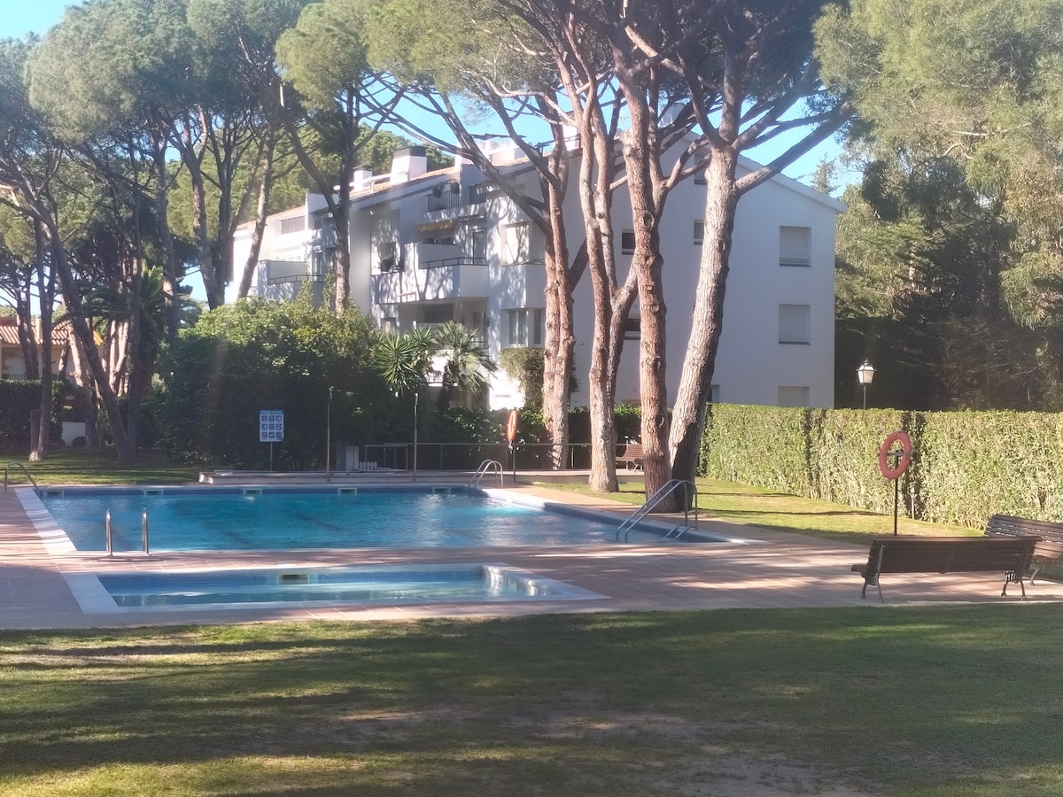 The outdoor area features a well-maintained swimming pool surrounded by lush green grass and tall trees. Lounge chairs are placed nearby, providing relaxation spots. The building in the background is a three-story structure, reflecting a calm residential environment.