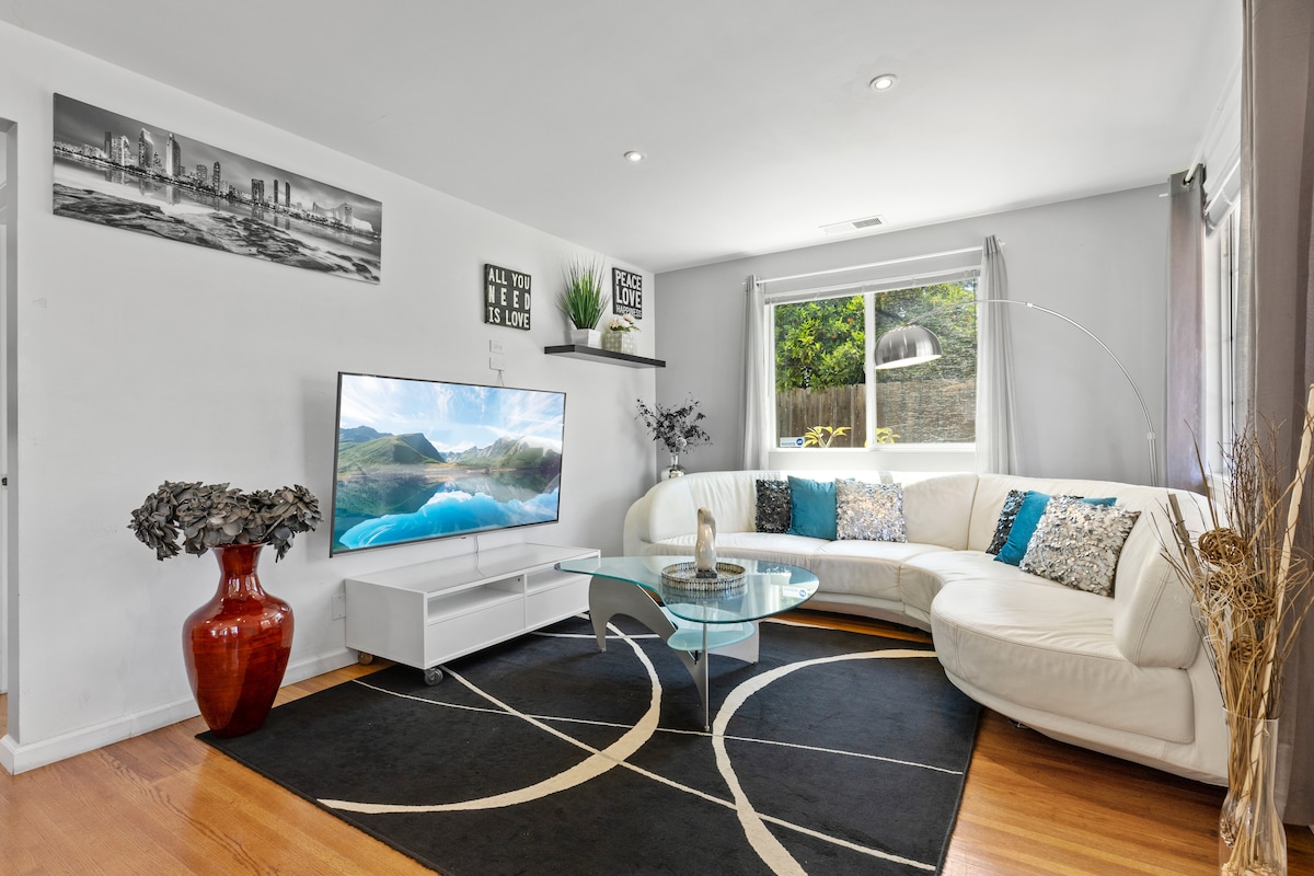 A bright living area features a large L-shaped white sofa adorned with colorful cushions. A modern glass coffee table sits on a patterned rug, and a large flat-screen TV is mounted on the wall. Natural light enters through the window, illuminating the space.